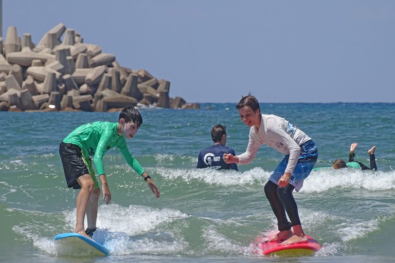 שני חברים חולקים גל יחד במהלך קייטנת גלישה לילדים בחופי הרצליה, נהנים מגלישת גלים משותפת."